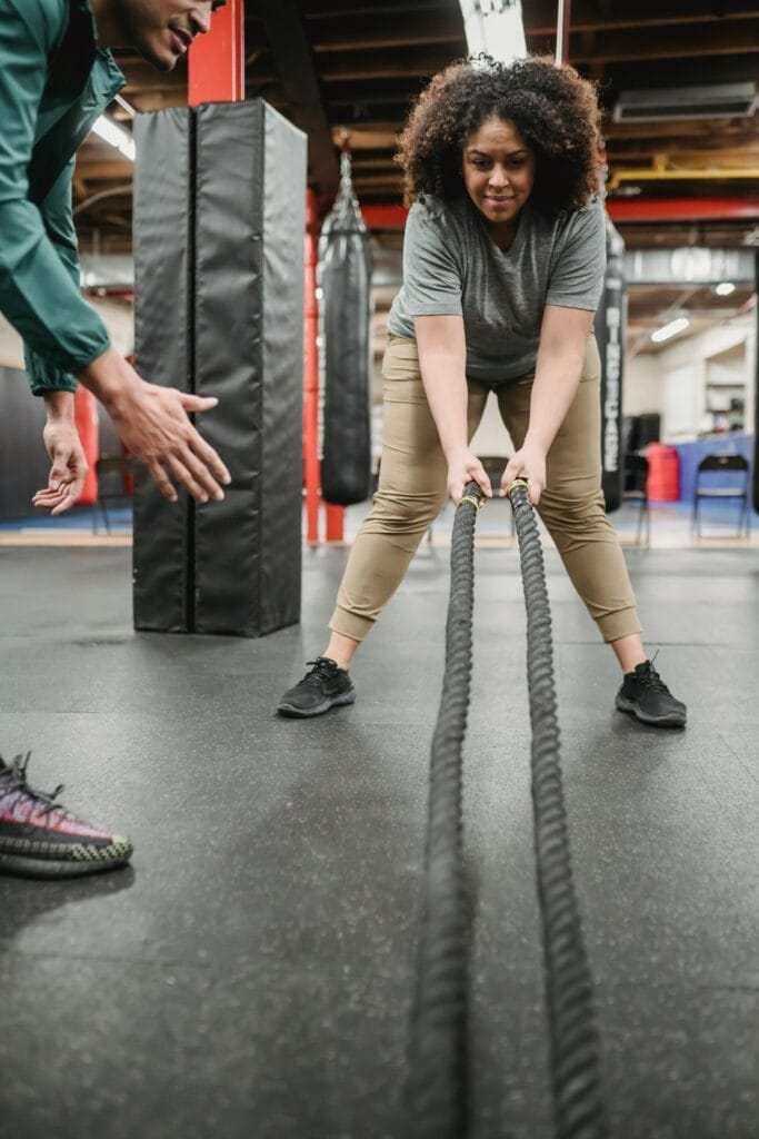 Positive plus sized African American female in sportswear with battle ropes listening to professional male trainer instructions in gym